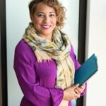 Smiling woman in a purple dress and scarf holding a tablet indoors.