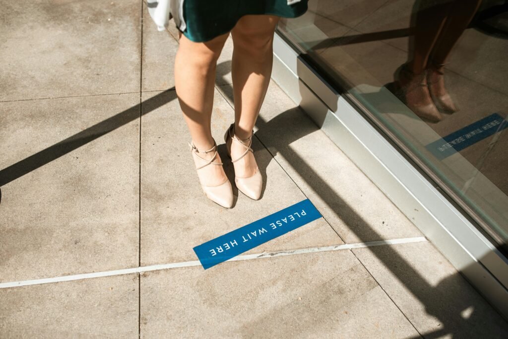 A person stands on a social distancing marker reading 'PLEASE WAIT HERE', shot outdoors.